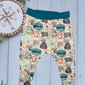 May include: Cream-colored baby pants with a teal waistband and cuffs. The pants feature a repeating pattern of camping-themed badges with bears, mountains, and campfires. The word "CAMP" is visible on some badges.