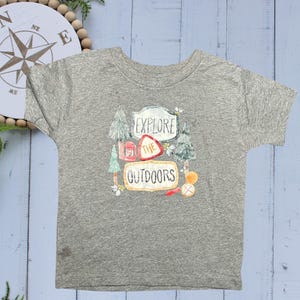 May include: Gray short-sleeved t-shirt with a colorful graphic that says "Explore the Outdoors." The graphic includes images of trees, a compass, and a campfire. The shirt is made of a soft, comfortable material.