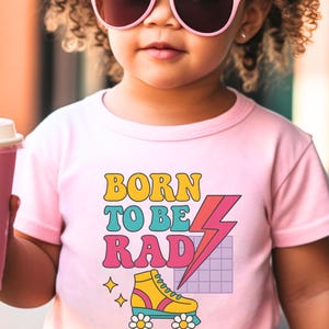 May include: A pink t-shirt with the text "BORN TO BE RAD" in yellow, teal, and pink. The shirt features a graphic of a yellow and teal roller skate with daisy wheels and a pink lightning bolt. The child is wearing pink sunglasses.
