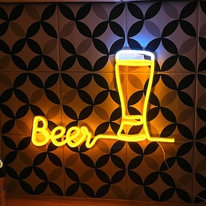 May include: A yellow neon sign featuring the word Beer in a cursive script next to a glowing outline of a beer glass. The sign emits a warm light against a patterned tile wall, creating a bright visual display of a beverage container.
