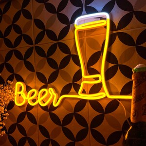 May include: A yellow neon sign displaying the word Beer in cursive script next to a glowing outline of a beer glass. The sign emits a warm light, illuminating the surrounding area with a bright yellow glow against a patterned tile wall.