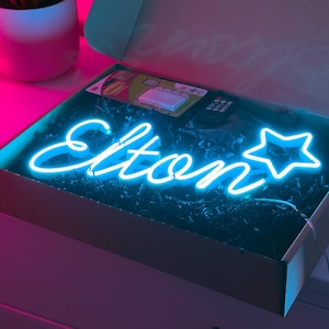 Peut inclure: Une enseigne néon bleu vif affichant le prénom Elton en écriture cursive, suivie d'une étoile à cinq branches. L'enseigne est placée dans une boîte en carton ouverte remplie de papier noir, accompagnée d'une petite télécommande et d'adhésifs.