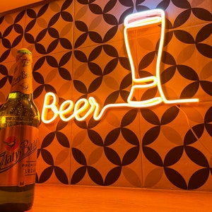 May include: A glowing yellow neon sign displaying the word Beer in cursive script next to an outline of a beer glass. Two glass beer bottles with labels reading Zlaty Bazant and Temny are positioned in the foreground of the illuminated wall sign.