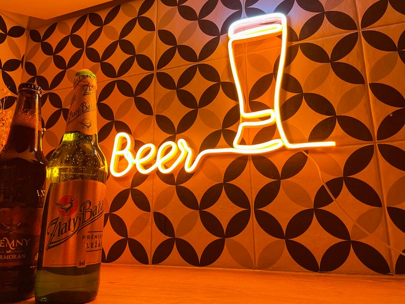 May include: A glowing yellow neon sign displaying the word Beer in cursive script next to an outline of a beer glass. Two glass beer bottles with labels reading Zlaty Bazant and Temny are positioned in the foreground of the illuminated wall sign.