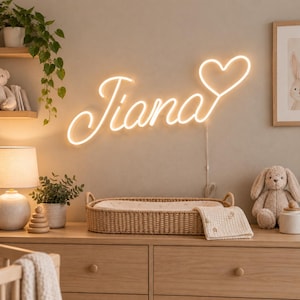 Peut inclure: Une enseigne néon LED blanc chaud fixée au mur, affichant le prénom Tiana dans une police cursive, se terminant par une forme de cœur. L'enseigne est composée d'un tube lumineux fin qui crée un éclairage doux sur la surface du mur.