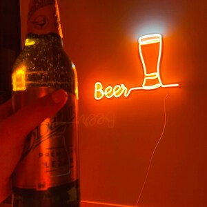 May include: A green glass beer bottle with a label featuring the text 'Pilsner Urquell' and 'Lezak' positioned in front of a wall-mounted orange neon sign that displays the word 'Beer' in cursive script next to a beer glass outline.