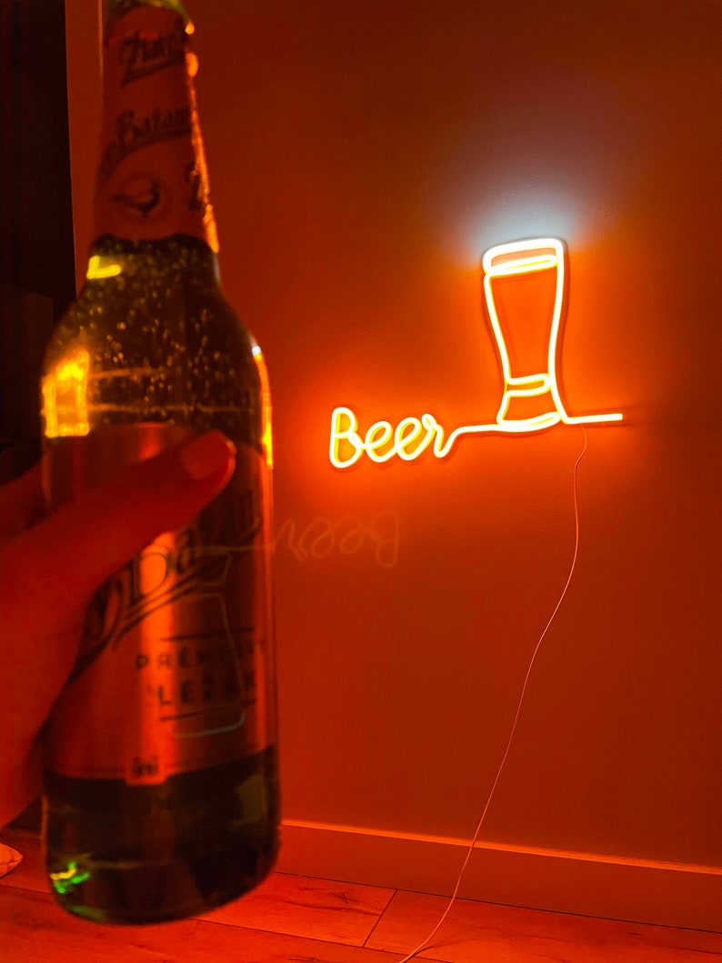 May include: A green glass beer bottle with a label featuring the text 'Pilsner Urquell' and 'Lezak' positioned in front of a wall-mounted orange neon sign that displays the word 'Beer' in cursive script next to a beer glass outline.