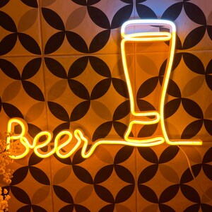 May include: A yellow neon sign featuring the word beer in a cursive script connected to the outline of a tall glass. The light emits a warm glow, creating a bright contrast against the dark lines of the wall behind the illuminated glass fixture.