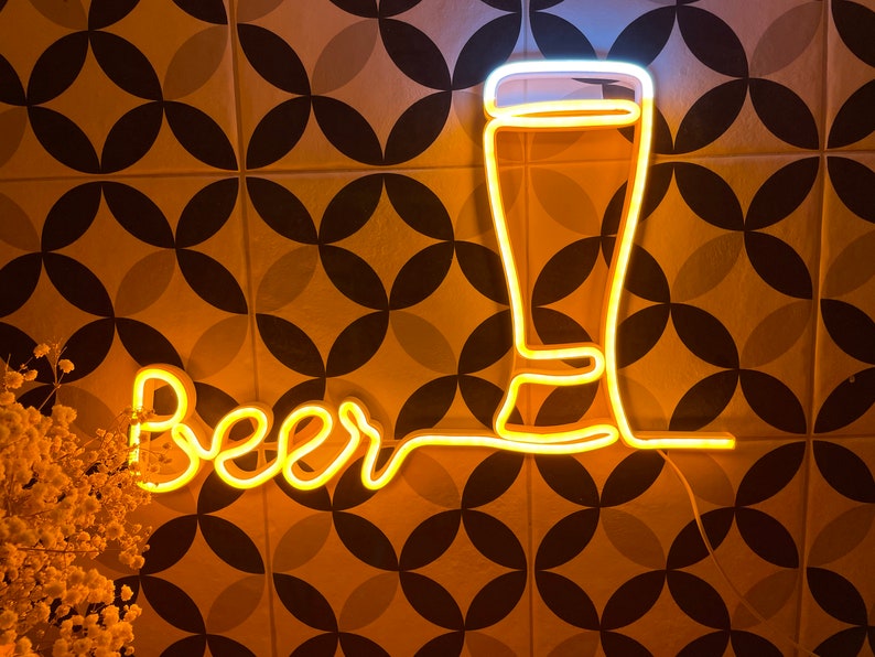 May include: A yellow neon sign featuring the word beer in a cursive script connected to the outline of a tall glass. The light emits a warm glow, creating a bright contrast against the dark lines of the wall behind the illuminated glass fixture.