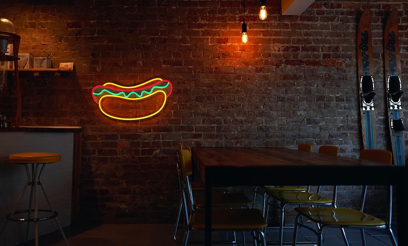 May include: A brick wall with a neon hot dog sign, a wooden table with yellow chairs, and two skis hanging on the wall.