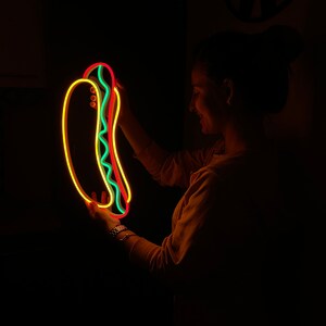 May include: A neon sign shaped like a hot dog with yellow, red, and green neon lights. The sign is being held by a person in a dark room.