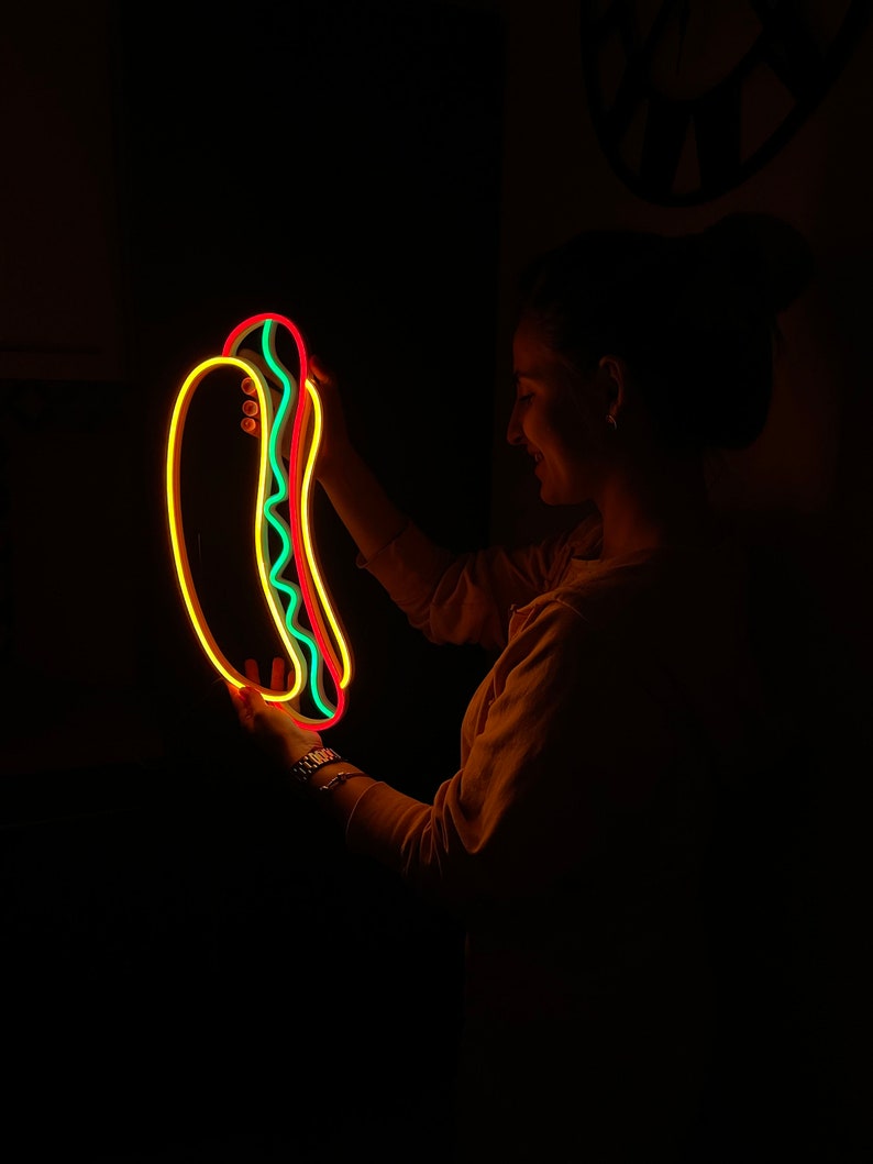 May include: A neon sign shaped like a hot dog with yellow, red, and green neon lights. The sign is being held by a person in a dark room.