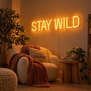 Peut inclure: Une enseigne néon fixée au mur émettant une lueur jaune chaude formant les mots STAY WILD dans une police sans empattement. L'enseigne est installée sur un mur texturé au-dessus d'un fauteuil crème recouvert d'un plaid texturé couleur rouille.