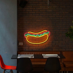 May include: Neon sign shaped like a hot dog with yellow mustard and green relish on a brick wall. The sign is hanging above a table with two chairs.