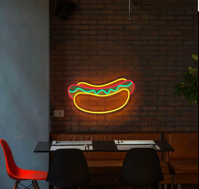 May include: Neon sign shaped like a hot dog with yellow mustard and green relish on a brick wall. The sign is hanging above a table with two chairs.