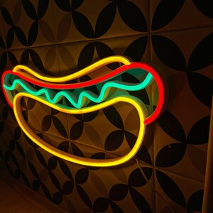 May include: A neon sign shaped like a hot dog with yellow, red, and blue neon lights. The sign is mounted on a wall with a patterned tile background.