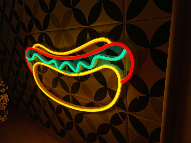 May include: A neon sign shaped like a hot dog with yellow, red, and blue neon lights. The sign is mounted on a wall with a patterned tile background.