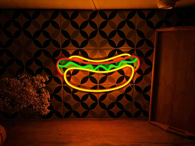 May include: A neon sign shaped like a hot dog with yellow mustard, green relish, and a red hot dog. The sign is lit up and glowing against a dark background.