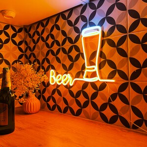 May include: A glowing yellow neon sign featuring the word Beer in a cursive script next to a simple outline of a tall glass. The sign is mounted on a wall with a geometric pattern of black and white circles. A dark glass bottle stands nearby.