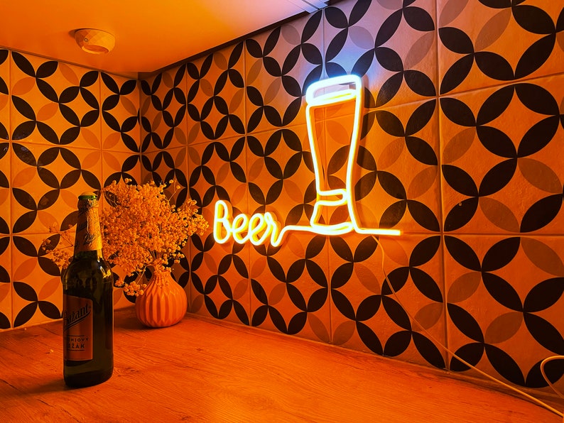 May include: A glowing yellow neon sign featuring the word Beer in a cursive script next to a simple outline of a tall glass. The sign is mounted on a wall with a geometric pattern of black and white circles. A dark glass bottle stands nearby.