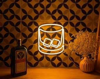 May include: A bright yellow neon sign shaped like a rocks glass containing two ice cubes. The sign is mounted on a wall with a geometric pattern of black and white circles. To the left is a bottle of Jägermeister and to the right is a vase of flowers.