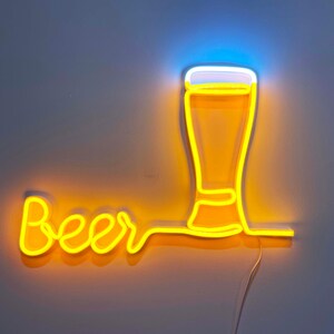 May include: A neon sign featuring the word Beer in yellow cursive script, connected to a glowing yellow outline of a beer glass with a white foam top. The sign emits a bright yellow and white light against a plain wall background.
