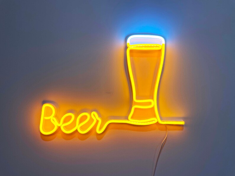 May include: A neon sign featuring the word Beer in yellow cursive script, connected to a glowing yellow outline of a beer glass with a white foam top. The sign emits a bright yellow and white light against a plain wall background.
