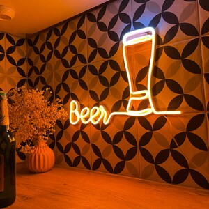 May include: A warm yellow neon sign displaying the word Beer in a cursive script next to a glowing outline of a tall beer glass. The sign emits a bright light, illuminating the surrounding area with a soft golden glow against a patterned wall.
