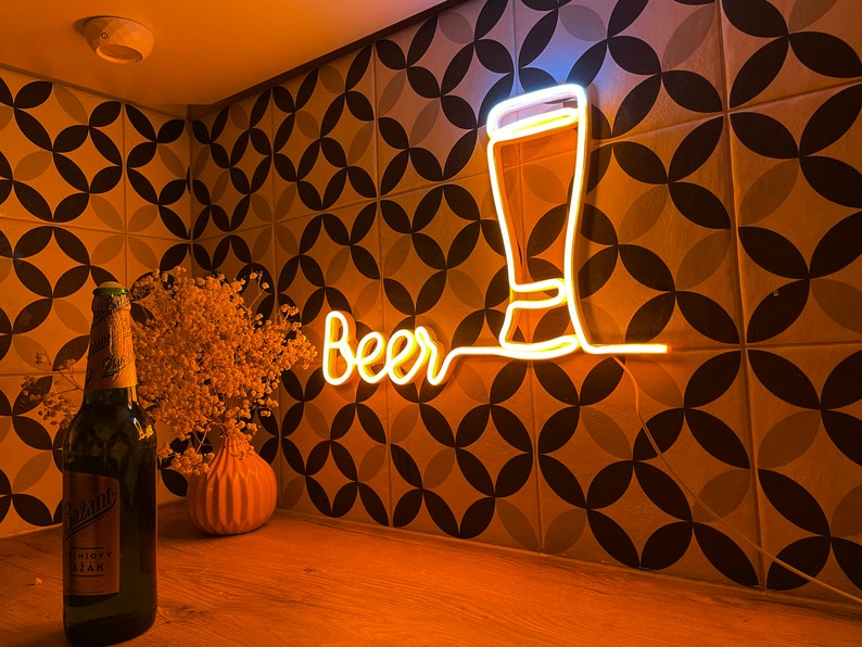 May include: A warm yellow neon sign displaying the word Beer in a cursive script next to a glowing outline of a tall beer glass. The sign emits a bright light, illuminating the surrounding area with a soft golden glow against a patterned wall.