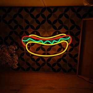 May include: A neon sign shaped like a hot dog with yellow, red, and blue accents. The sign is mounted on a wall with a brown and green patterned tile background.