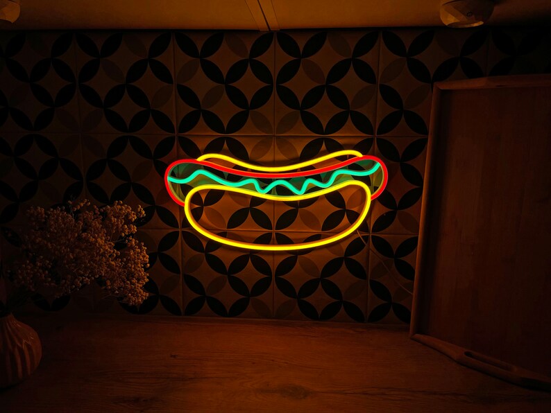 May include: A neon sign shaped like a hot dog with yellow, red, and blue accents. The sign is mounted on a wall with a brown and green patterned tile background.