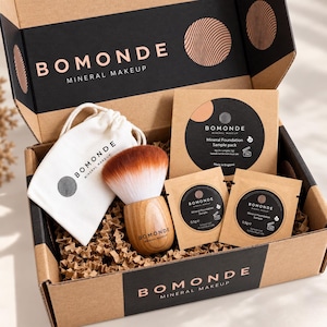 May include: A Bomonde Mineral Makeup box containing a white drawstring pouch, a makeup brush with a wooden handle and brown-tipped bristles, a square foundation sample pack, and two small individual foundation sample sachets, all on brown paper.