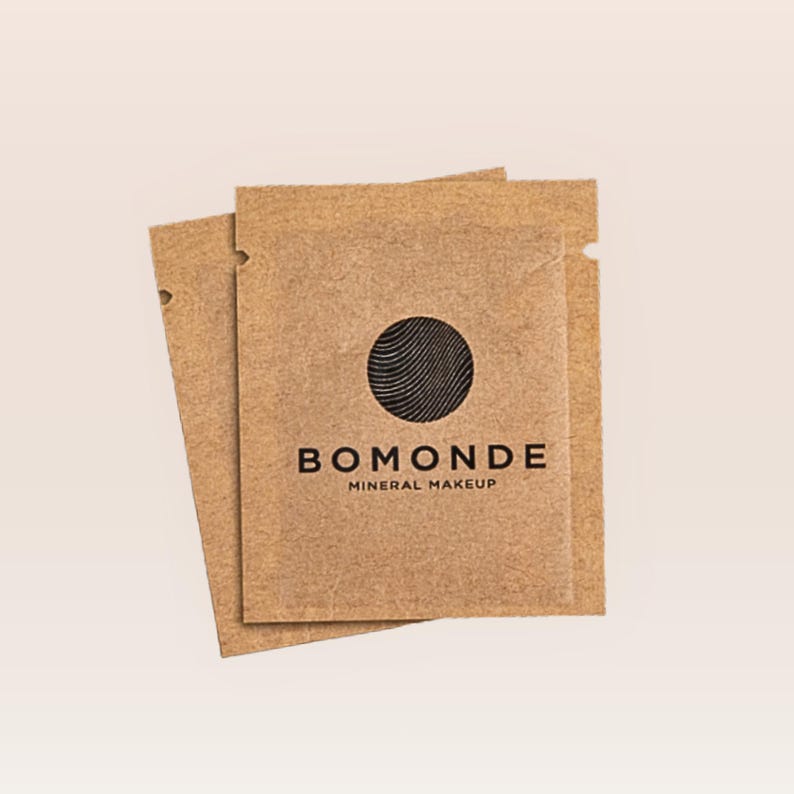 May include: Two small, brown paper packets with the text "BOMONDE MINERAL MAKEUP" in black. Each packet has a black circle with curved lines. The packets are slightly overlapping, with one angled to the left.