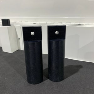 May include: Two black plastic cylindrical pieces with a square top and a hole in the center. The pieces are on a black surface.