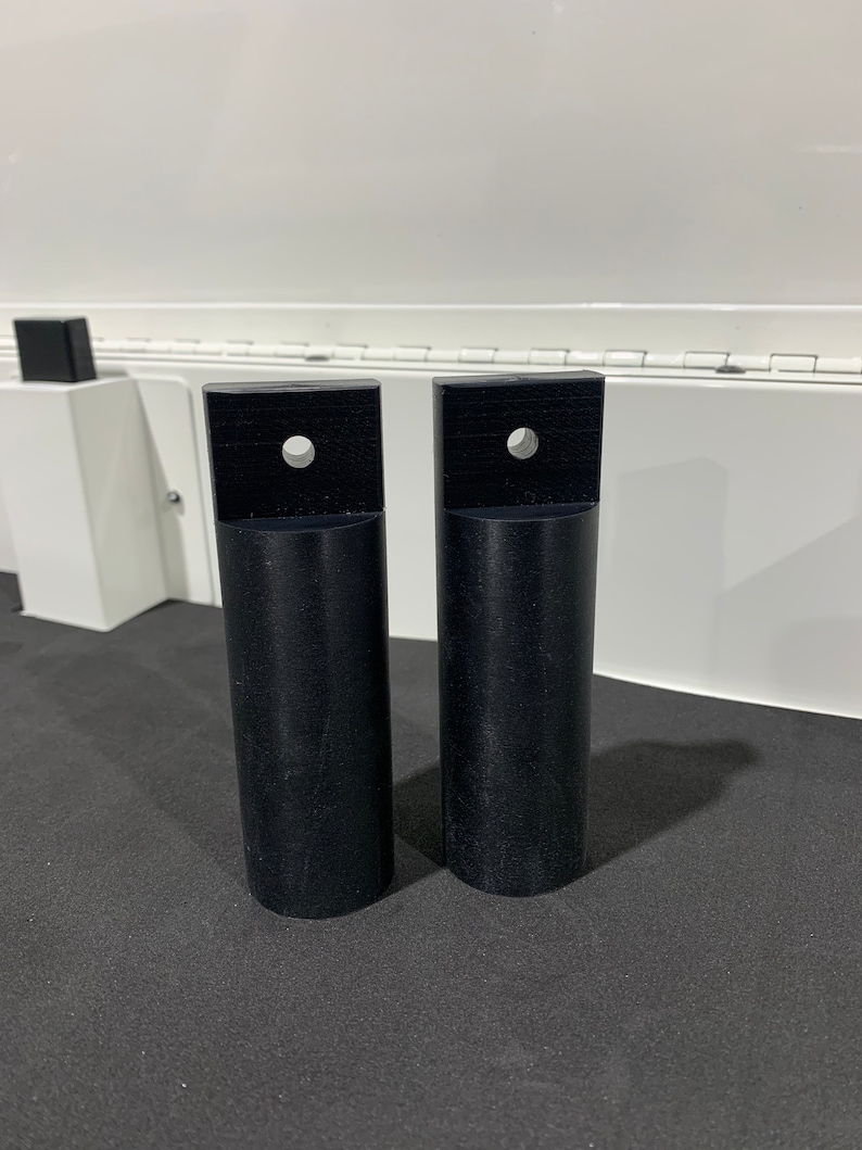 May include: Two black plastic cylindrical pieces with a square top and a hole in the center. The pieces are on a black surface.