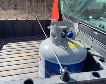 May include: A gray propane tank with blue accents and a white hose is secured in the bed of a utility vehicle. An orange traffic cone is visible near the windshield. The background shows a sunny outdoor scene with trees.