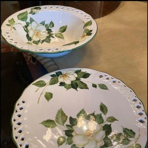 Puede incluir: Un juego de dos cuencos de cerámica blanca con ribete verde y diseños florales. Los cuencos tienen un borde festoneado y un delicado diseño calado alrededor del borde.