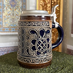 Könnte beinhalten: Ein Bierkrug aus Keramik mit dunkelbrauner und blauer Glasur sowie geprägten Blumen- und geometrischen Mustern. Das Gefäß verfügt über einen metallischen Klappdeckel und einen stabilen Henkel, die detaillierte Reliefverzierungen aufweisen.