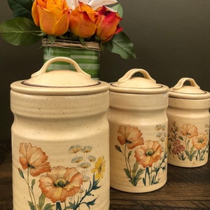 Puede incluir: Tres botes de cerámica con diseños florales. Los botes son de color beige con detalles marrones y presentan un patrón floral de amapolas naranjas y hojas verdes. Los botes están dispuestos en fila sobre una superficie de madera.