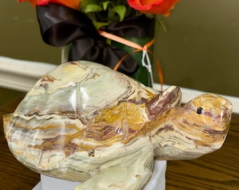 May include: A carved stone turtle figurine with a multi-colored pattern of green, brown, and yellow. The turtle is resting on a white surface, with a vase of orange roses and a yellow sunflower in the background.