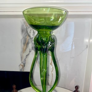 May include: A tall, green glass decorative vase with a wide, shallow bowl at the top. The vase has a unique, elongated design with curved, petal-like supports. The base is circular with concentric green rings. Two small brown chess pieces are on the table.