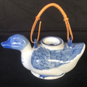 Puede incluir: Una tetera de porcelana azul y blanca con forma de pato y un asa de mimbre trenzado. La tetera tiene un diseño floral azul y blanco.