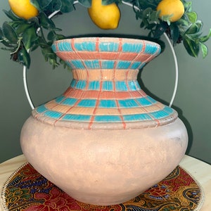 Puede incluir: Un jarrón de terracota con un patrón geométrico en tonos de rosa, azul y blanco. El jarrón tiene una base ancha y un cuello estrecho.