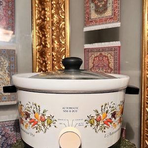 Puede incluir: Olla de cocción lenta Automatic Sim-R-Pot blanca y negra con tapa de cristal y asas negras. La olla de cocción lenta tiene un diseño floral con verduras y el texto "Automatic Sim-R-Pot".