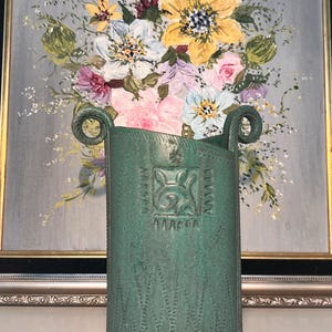 Pode incluir: Um vaso cilíndrico de cerâmica verde com superfície texturizada, apresentando duas alças enroladas no topo e um design central em relevo quadrado. O vaso tem um acabamento fosco e é decorado com padrões de pontos verticais na base.