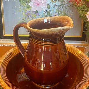 Może przedstawiać: Wazon z brązowej ceramiki w stylu vintage z pasującą miską. Dzbanek ma szeroki dzióbek i zakrzywiony uchwyt. Misa ma szeroką krawędź z teksturowanym wzorem. Zestaw ma ciepły brązowy kolor z błyszczącym wykończeniem.