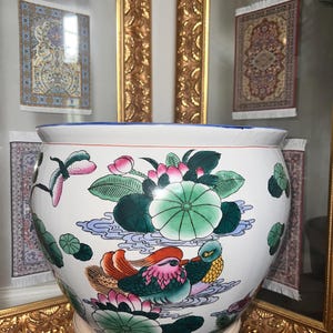 Puede incluir: Una gran maceta de cerámica blanca con un borde azul y un colorido diseño floral y de aves. El diseño incluye flores de loto rosas, hojas de nenúfar verdes y dos patos mandarines. La maceta se apoya en un soporte blanco.