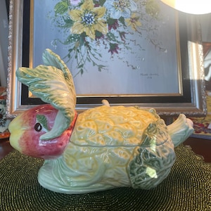 Puede incluir: Un recipiente de cerámica con forma de manzana con una combinación de colores amarillo, verde y rojo. El recipiente tiene tapa y está diseñado para parecerse a una manzana. El fondo incluye una pintura de flores.