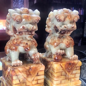 May include: A pair of jade foo dog statues with intricate details, each standing on a square base with a brick pattern. The statues are a light green color with brown accents.