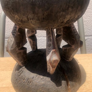 Puede incluir: Un taburete de madera con un cuenco tallado en la parte superior y cuatro patas talladas de forma intrincada. El taburete es de color marrón oscuro con toques de pintura roja y blanca.